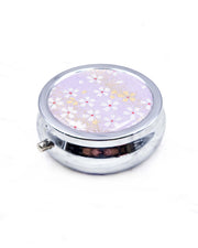 Round silver metal pill box featuring small white and gold cherry blossoms on a purple background with metallic gold details.