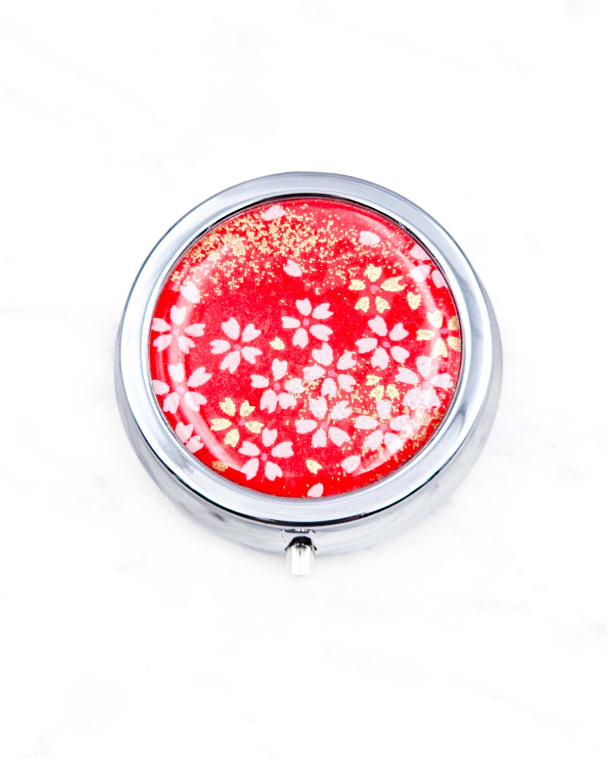 Round silver metal pill box featuring small white and gold cherry blossoms on a red background with metallic gold details.