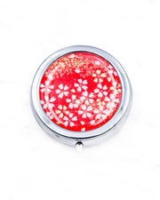 Round silver metal pill box featuring small white and gold cherry blossoms on a red background with metallic gold details.