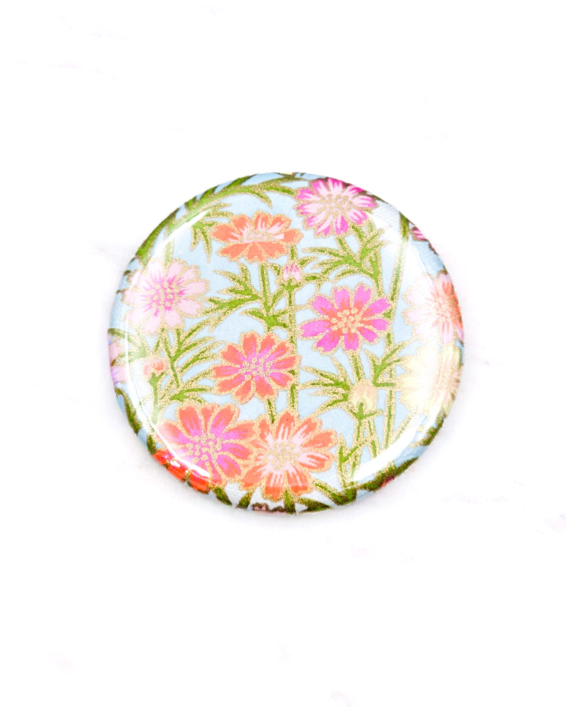 Round pocket mirror featuring pink and orange flower stalks on a light blue background with metallic gold accents.