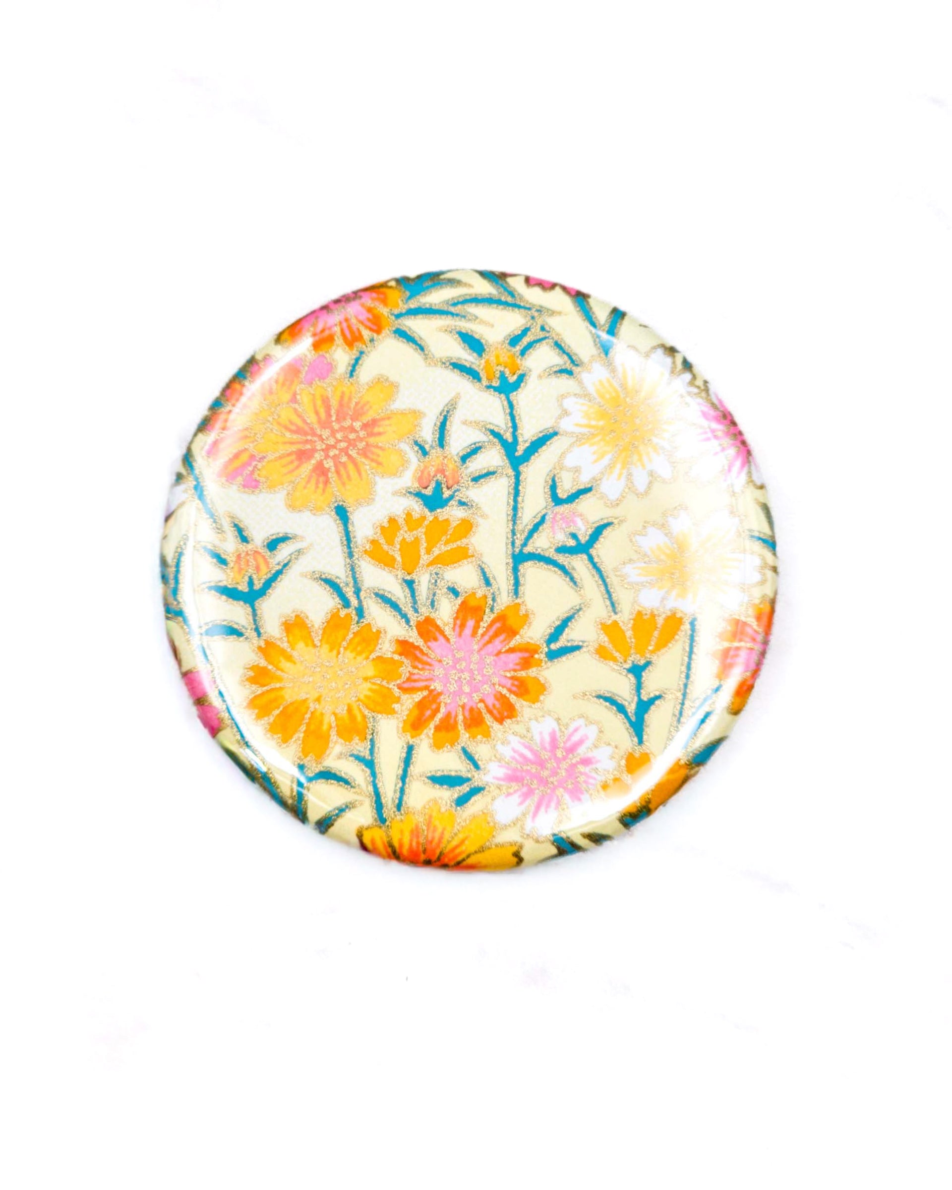 Round pocket mirror featuring orange and white flower stalks on a beige background with metallic gold accents.