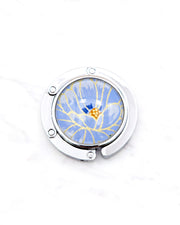 Round silver metal table-top bag hanger featuring a large blue chrysanthemum flower with metallic gold details.