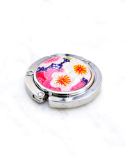 Round silver metal table-top bag hanger featuring pink and white cherry blossoms on a dark purple background with metallic gold details.