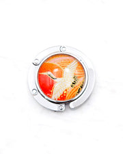 Round silver metal table-top bag hanger featuring a white crane on an orange background with metallic gold details.