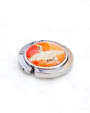 Round silver metal table-top bag hanger featuring a white crane on an orange background with metallic gold details.