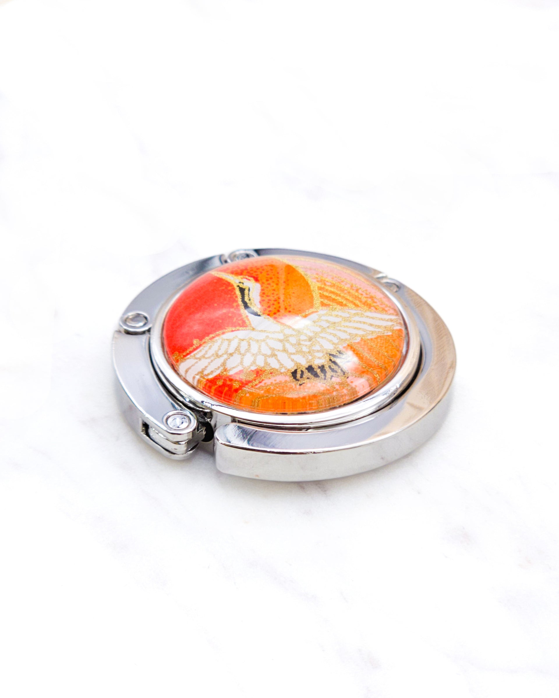 Round silver metal table-top bag hanger featuring a white crane on an orange background with metallic gold details.