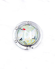 Round silver metal tabletop bag hanger featuring a white flying crane on a turquoise green background with metallic gold details.