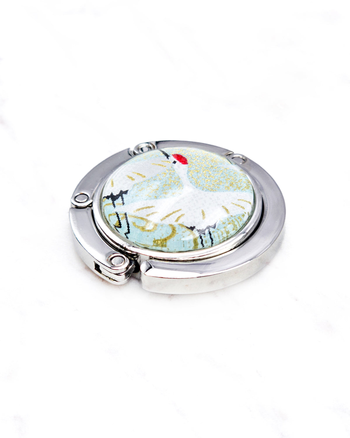 Round silver metal tabletop bag hanger featuring a white flying crane on a turquoise green background with metallic gold details.
