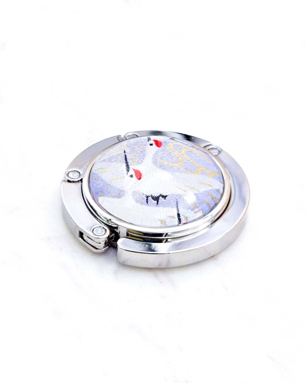 Round silver metal table-top bag hanger featuring flying white cranes on a light purple background with metallic gold details.