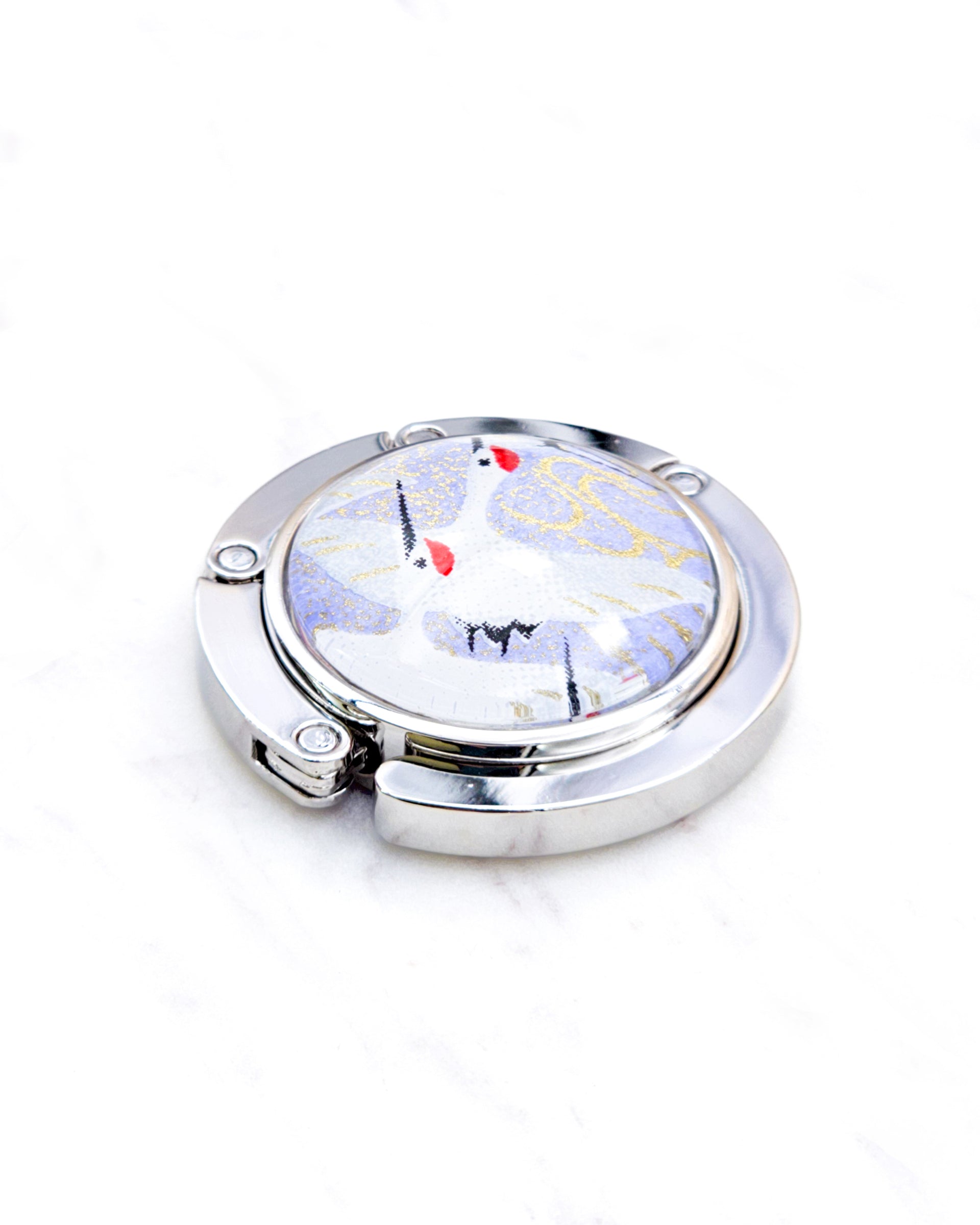 Round silver metal table-top bag hanger featuring flying white cranes on a light purple background with metallic gold details.