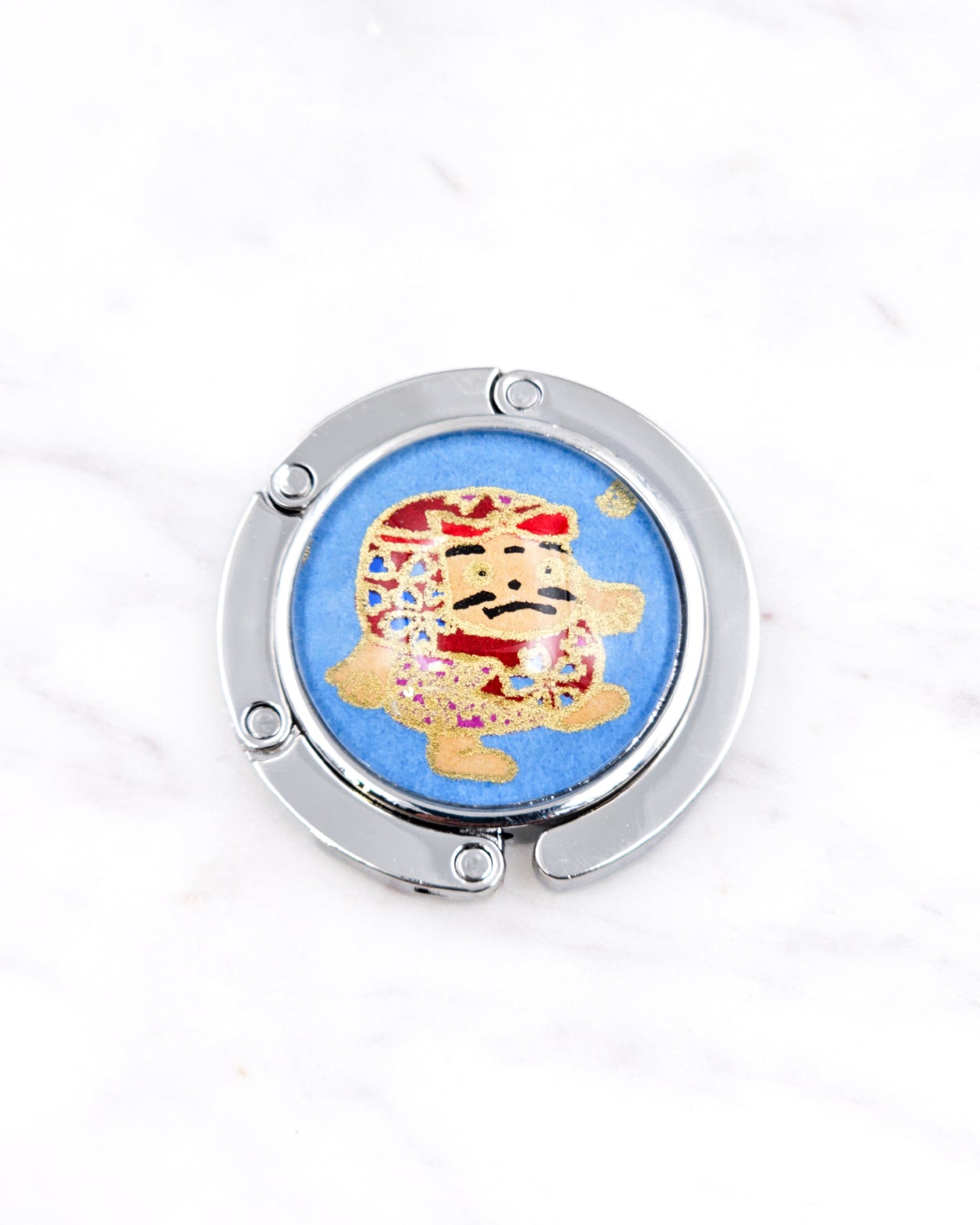 Silver metal table-top bag hanger featuring a red daruma doll on a blue background with metallic gold accents.