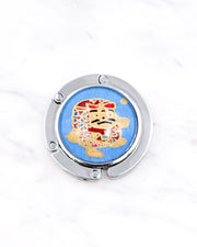 Silver metal table-top bag hanger featuring a red daruma doll on a blue background with metallic gold accents.