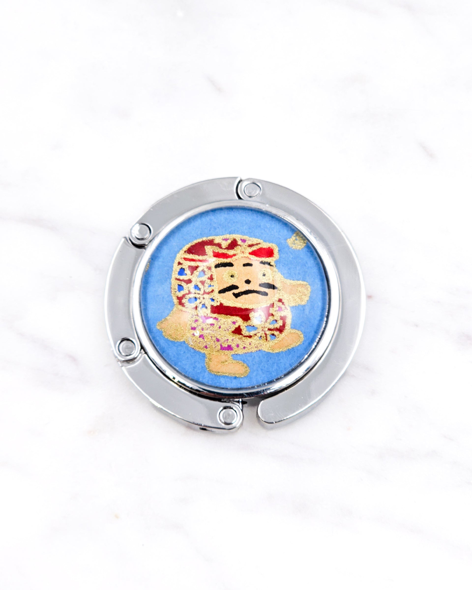 Silver metal table-top bag hanger featuring a red daruma doll on a blue background with metallic gold accents.