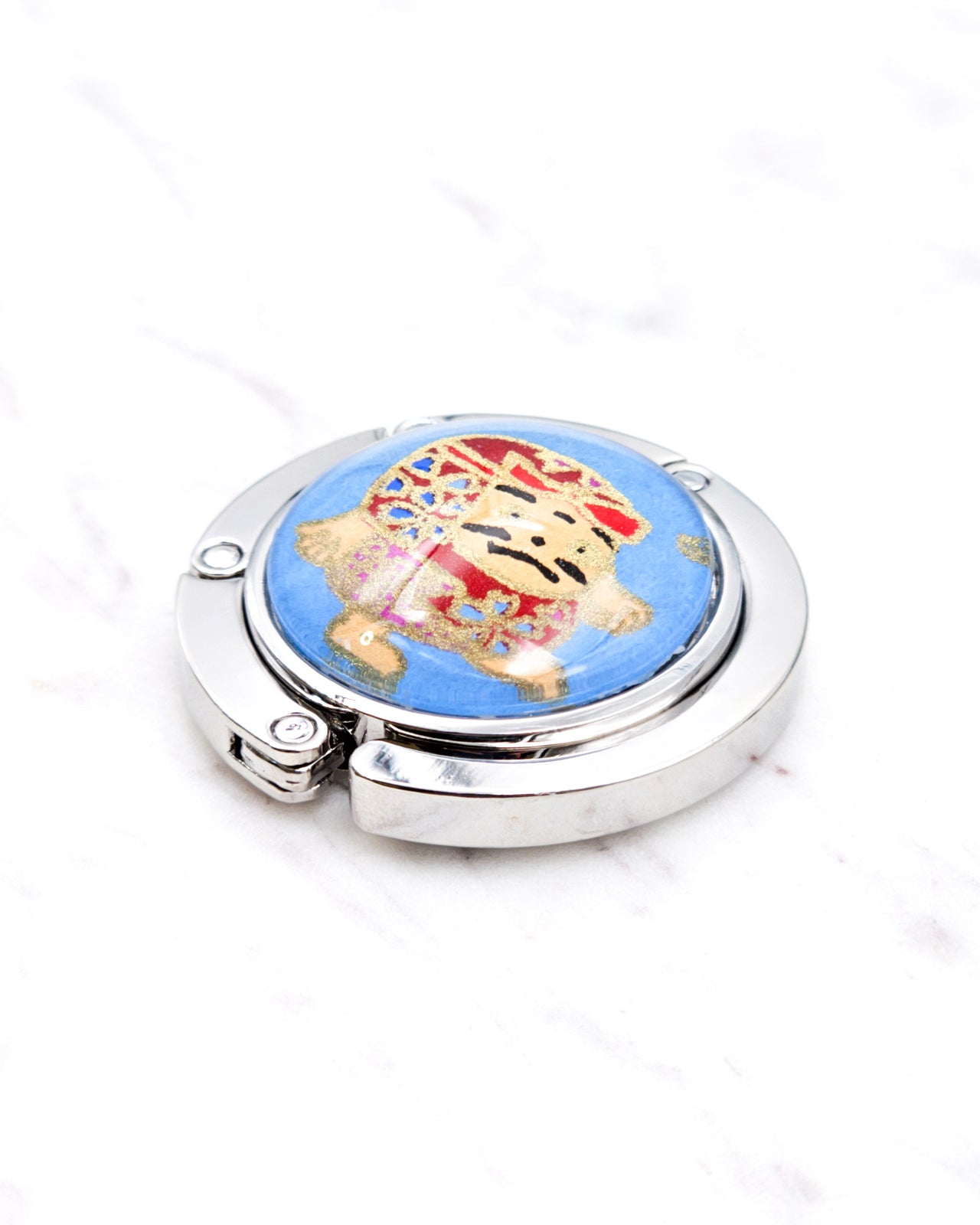 Silver metal table-top bag hanger featuring a red daruma doll on a blue background with metallic gold accents.