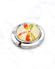 Silver metal table-top bag hanger featuring a red and green dragonfly on a yellow background with metallic gold accents.