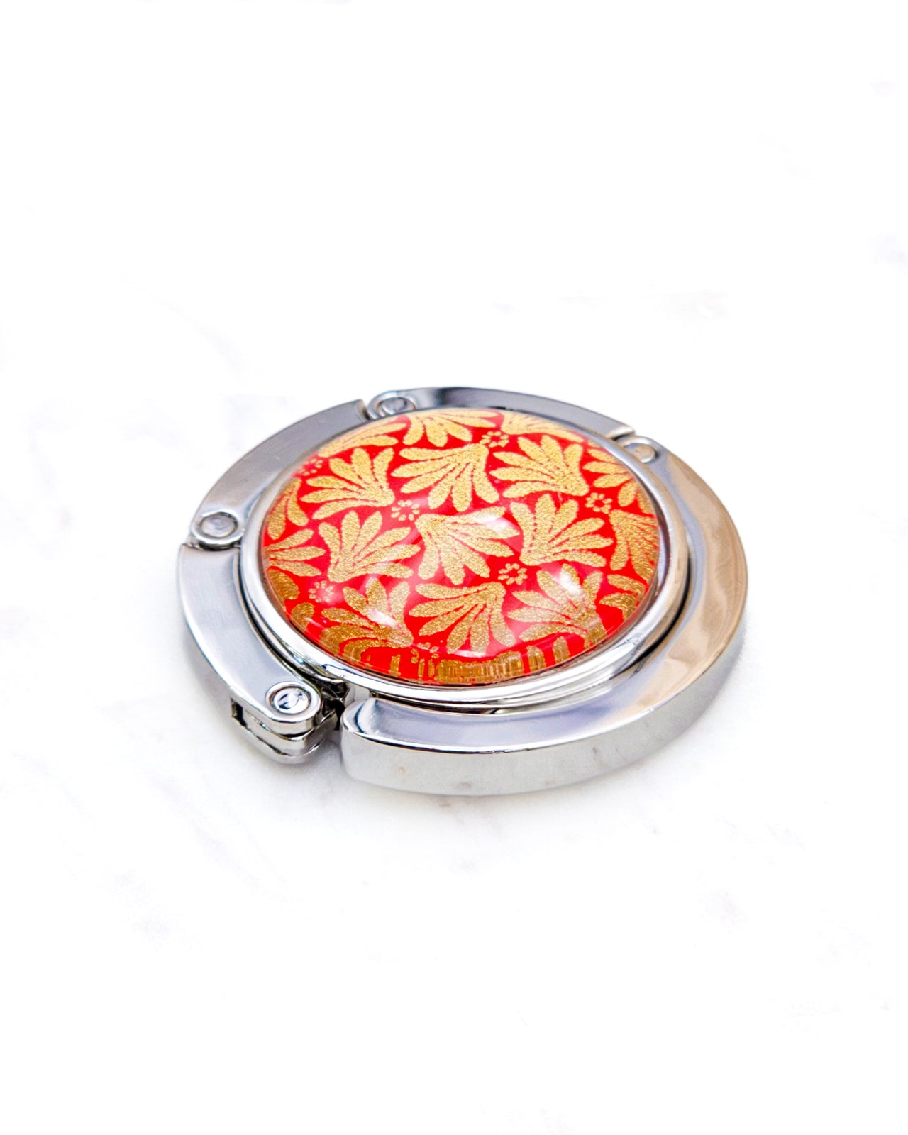 Round silver metal table-top bag hanger featuring a metallic gold motif on a red background.