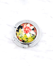 Silver metal table-top bag hanger featuring green, red, and white maples leaves on a black background with metallic gold accents.