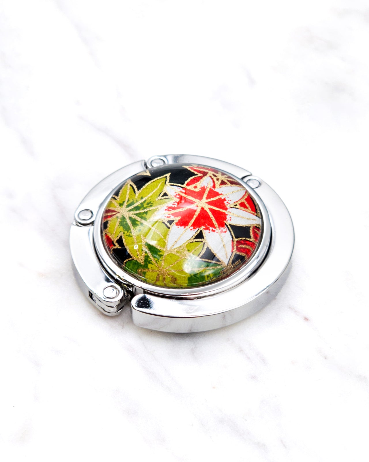 Silver metal table-top bag hanger featuring green, red, and white maples leaves on a black background with metallic gold accents.