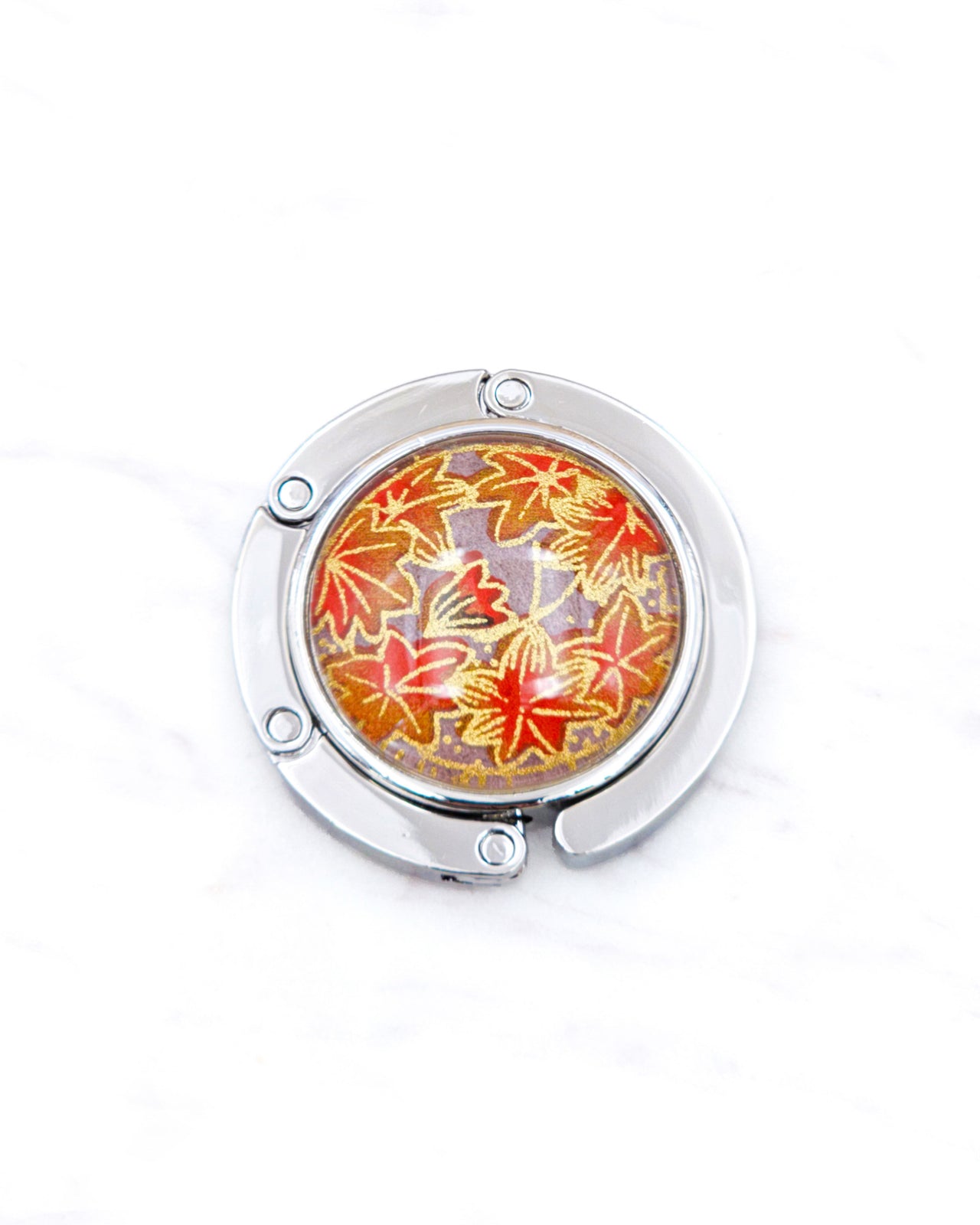 Round silver metal table-top bag hanger featuring orange maple leaves on a brown background with metallic gold details.