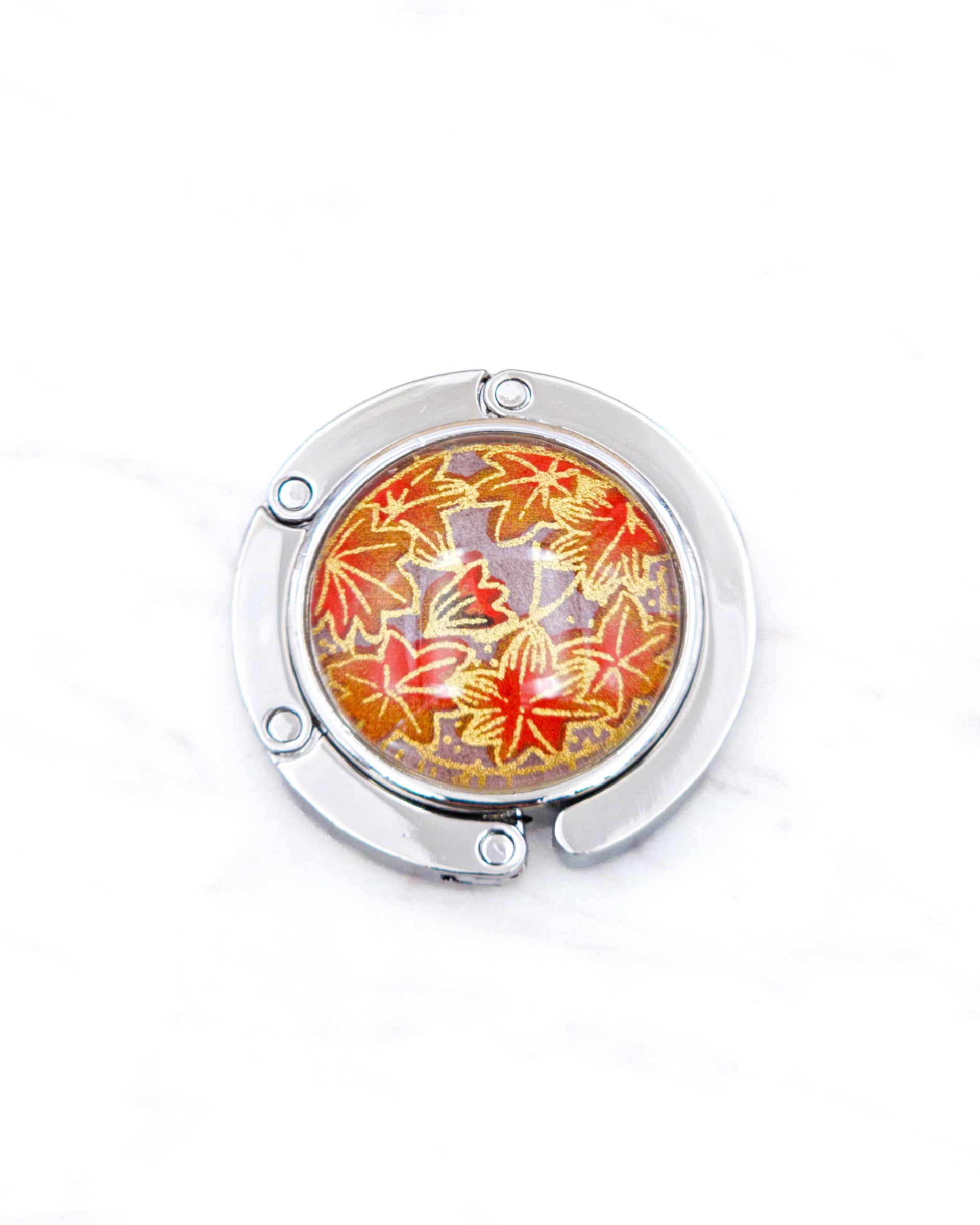 Round silver metal table-top bag hanger featuring orange maple leaves on a brown background with metallic gold details.