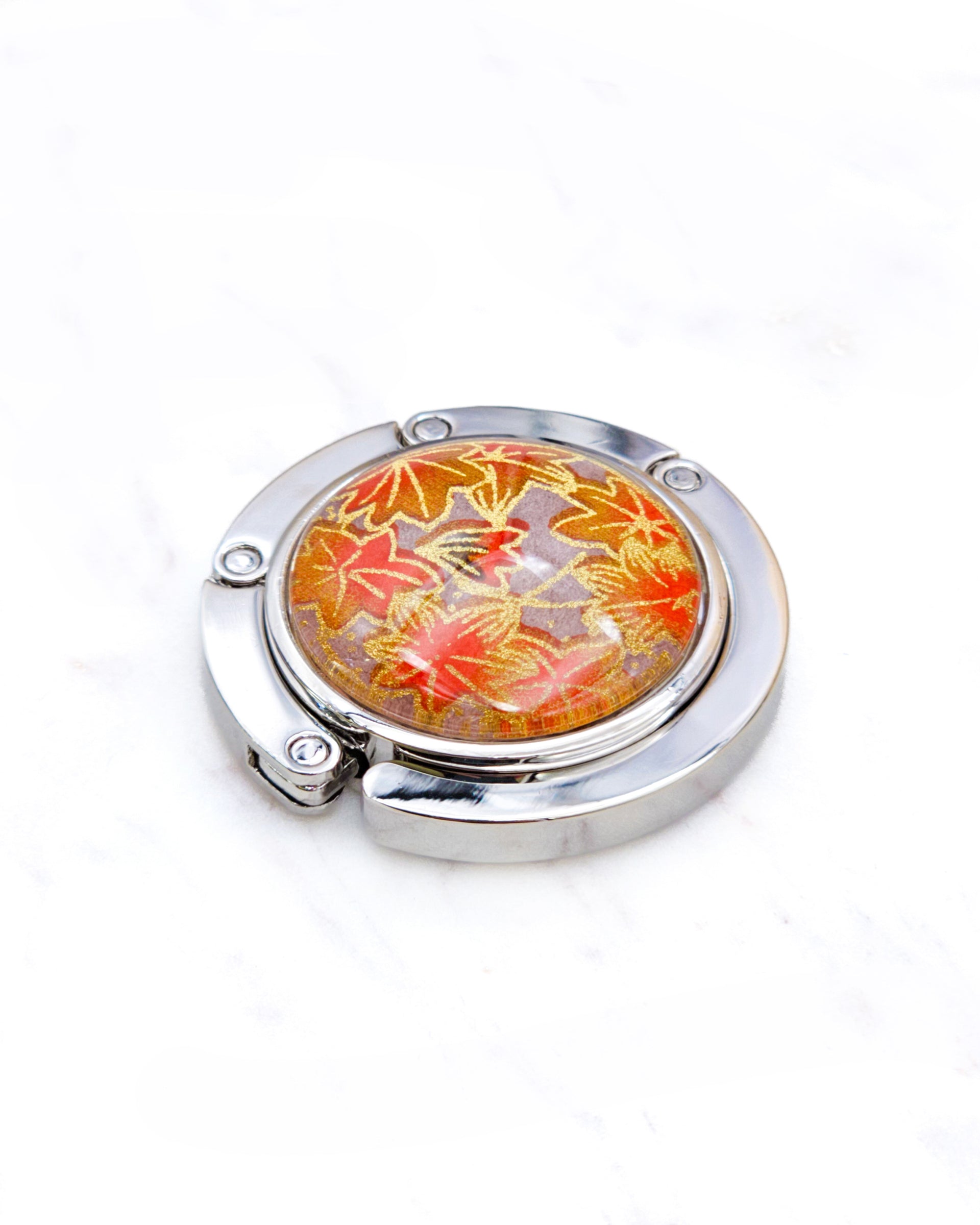 Round silver metal table-top bag hanger featuring orange maple leaves on a brown background with metallic gold details.