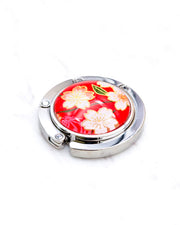 Round silver metal table-top bag hanger featuring pink and white cherry blossoms on a red background with metallic gold details.