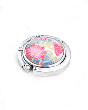 Round silver metal tabletop bag hanger featuring pink flowers with green stems on a blue and white background with metallic gold details.
