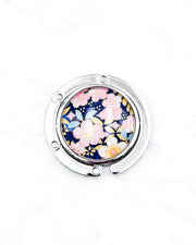 Round silver metal tabletop bag hanger featuring pink plum blossoms on a dark blue background with metallic gold details.