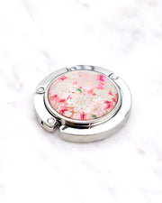 Silver metal table-top bag hanger featuring pink and white cherry blossoms on a pink background with metallic gold accents.