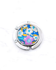 Round silver metal table-top bag hanger featuring pink and white cherry blossoms on a blue background with metallic gold details.