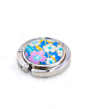 Round silver metal table-top bag hanger featuring pink and white cherry blossoms on a blue background with metallic gold details.