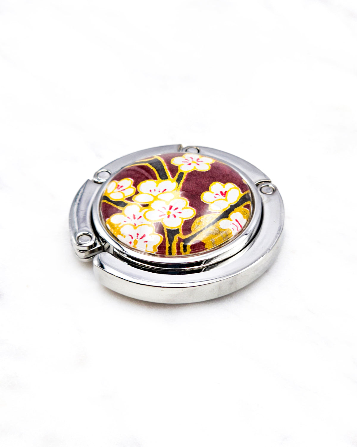 Round silver metal tabletop bag hanger featuring white plum blossoms and black branches on a brown background with metallic gold details.