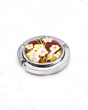 Round silver metal tabletop bag hanger featuring white plum blossoms and black branches on a brown background with metallic gold details.