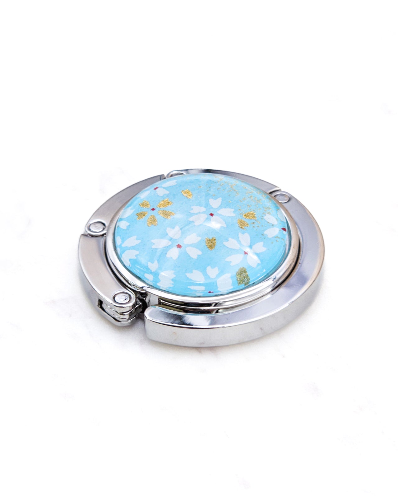 Round silver metal table-top bag hanger featuring small white and gold cherry blossoms on an aqua blue background.