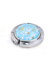 Round silver metal table-top bag hanger featuring small white and gold cherry blossoms on an aqua blue background.