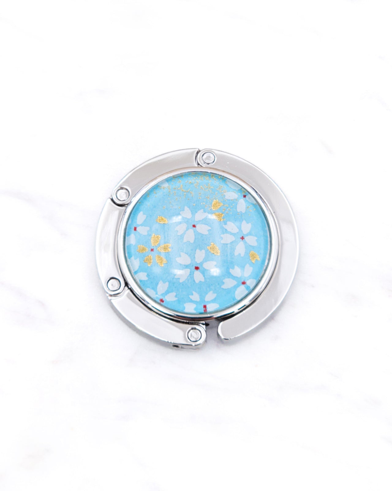 Round silver metal table-top bag hanger featuring small white and gold cherry blossoms on an aqua blue background.