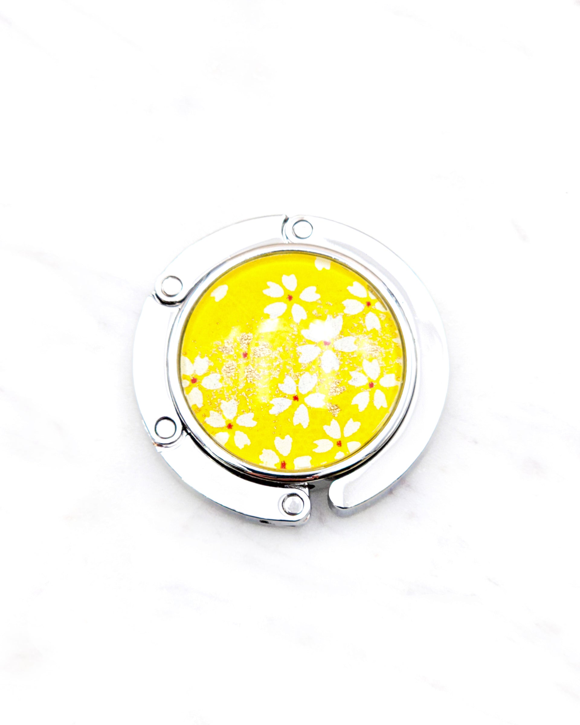 Round silver metal table-top bag hanger featuring small white and gold cherry blossoms on a bright yellow background with metallic gold details.