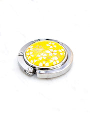 Round silver metal table-top bag hanger featuring small white and gold cherry blossoms on a bright yellow background with metallic gold details.