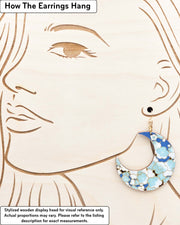 X-Large wooden Crescent Moon shaped earrings featuring turquoise blue and white plum blossom tree with dark brown branches on a blue background with metallic gold details, displayed on a stylized wooden head to demonstrate how the earring hangs when worn. Display is not to scale.