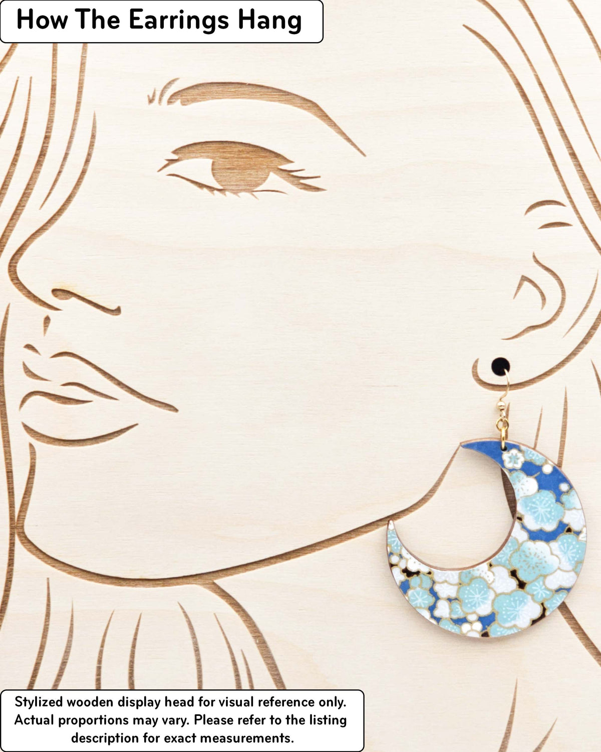 X-Large wooden Crescent Moon shaped earrings featuring turquoise blue and white plum blossom tree with dark brown branches on a blue background with metallic gold details, displayed on a stylized wooden head to demonstrate how the earring hangs when worn. Display is not to scale.