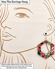 X-Large wooden hollow hexagon shaped earrings featuring red, green, and white maples leaves on a black background with metallic gold details, displayed on a stylized wooden head to demonstrate how the earring hangs when worn. Display is not to scale.