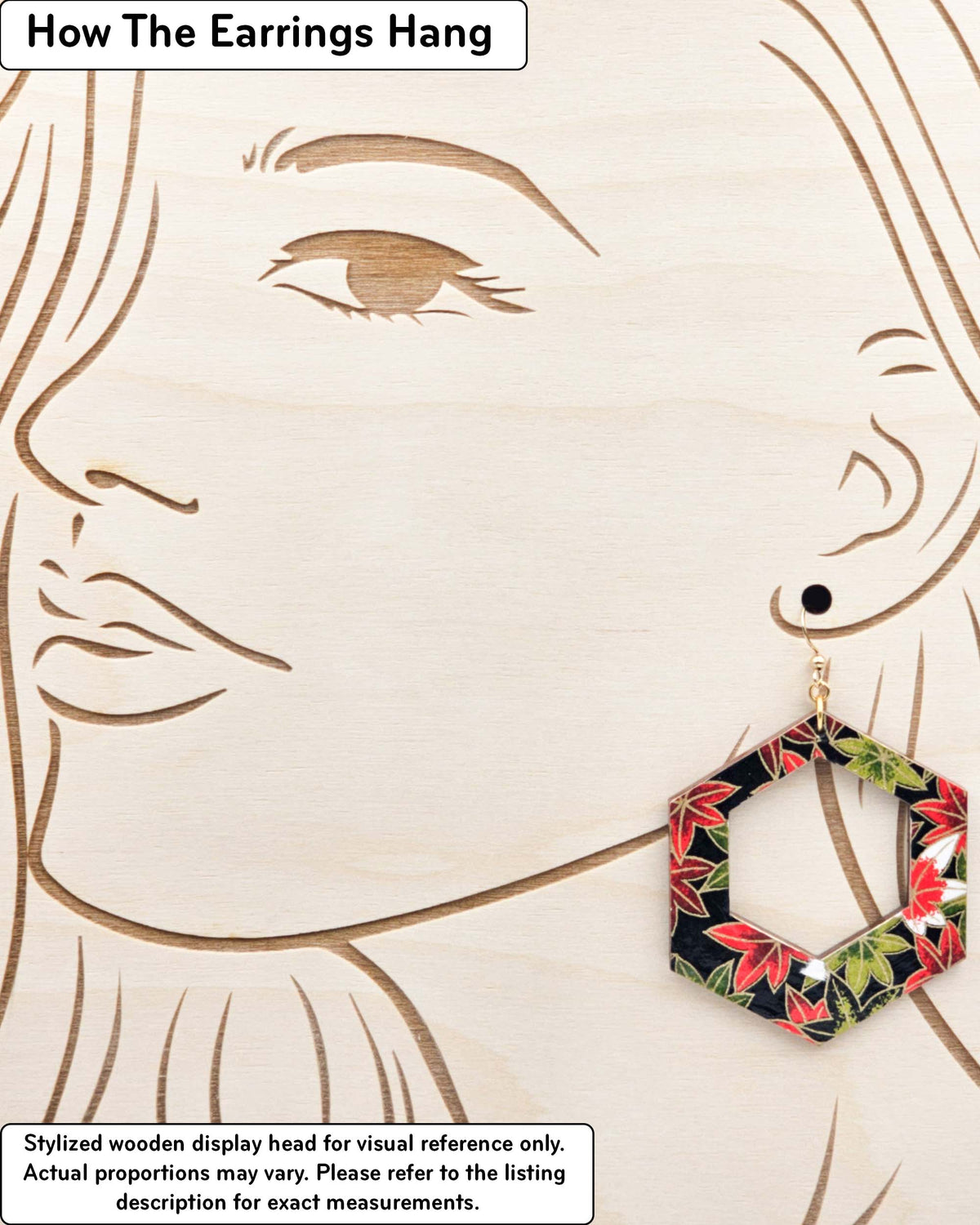 X-Large wooden hollow hexagon shaped earrings featuring red, green, and white maples leaves on a black background with metallic gold details, displayed on a stylized wooden head to demonstrate how the earring hangs when worn. Display is not to scale.