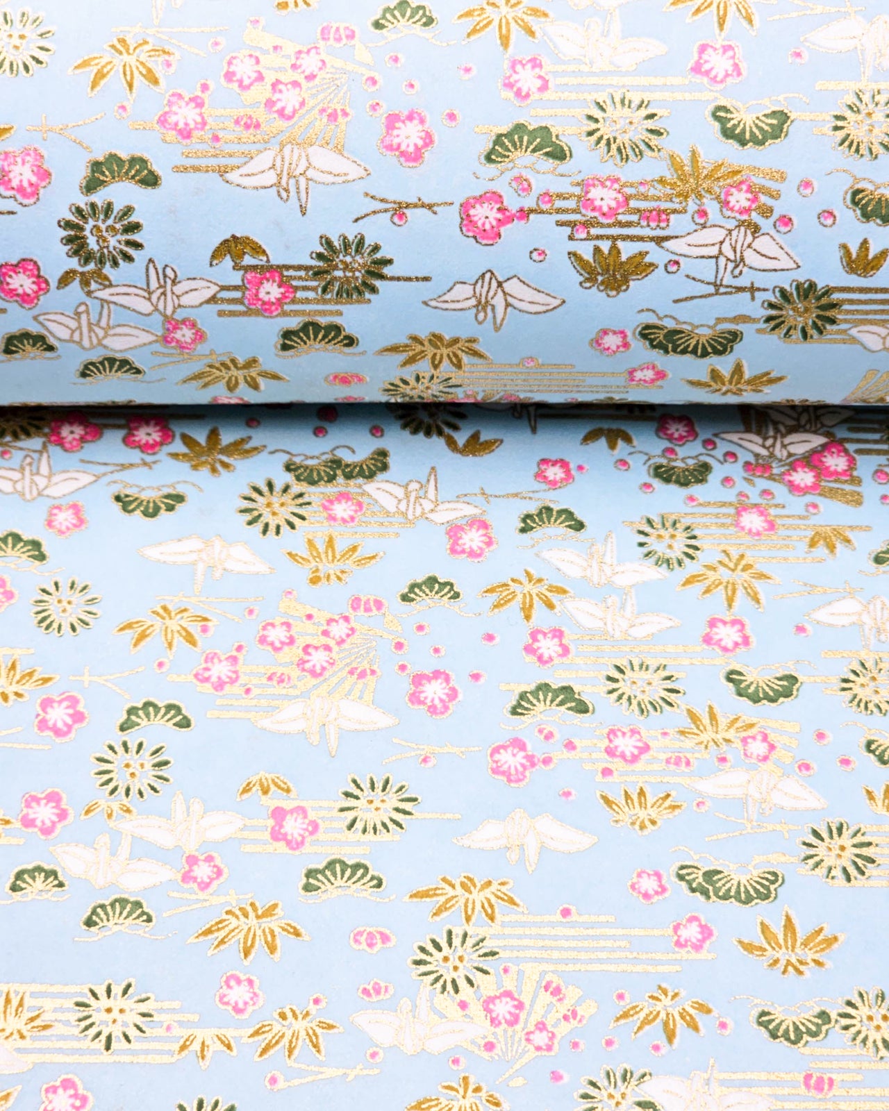 Japanese chiyogami paper featuring white origami cranes with green plants and pink plum blossoms on a light blue background with metallic gold details.