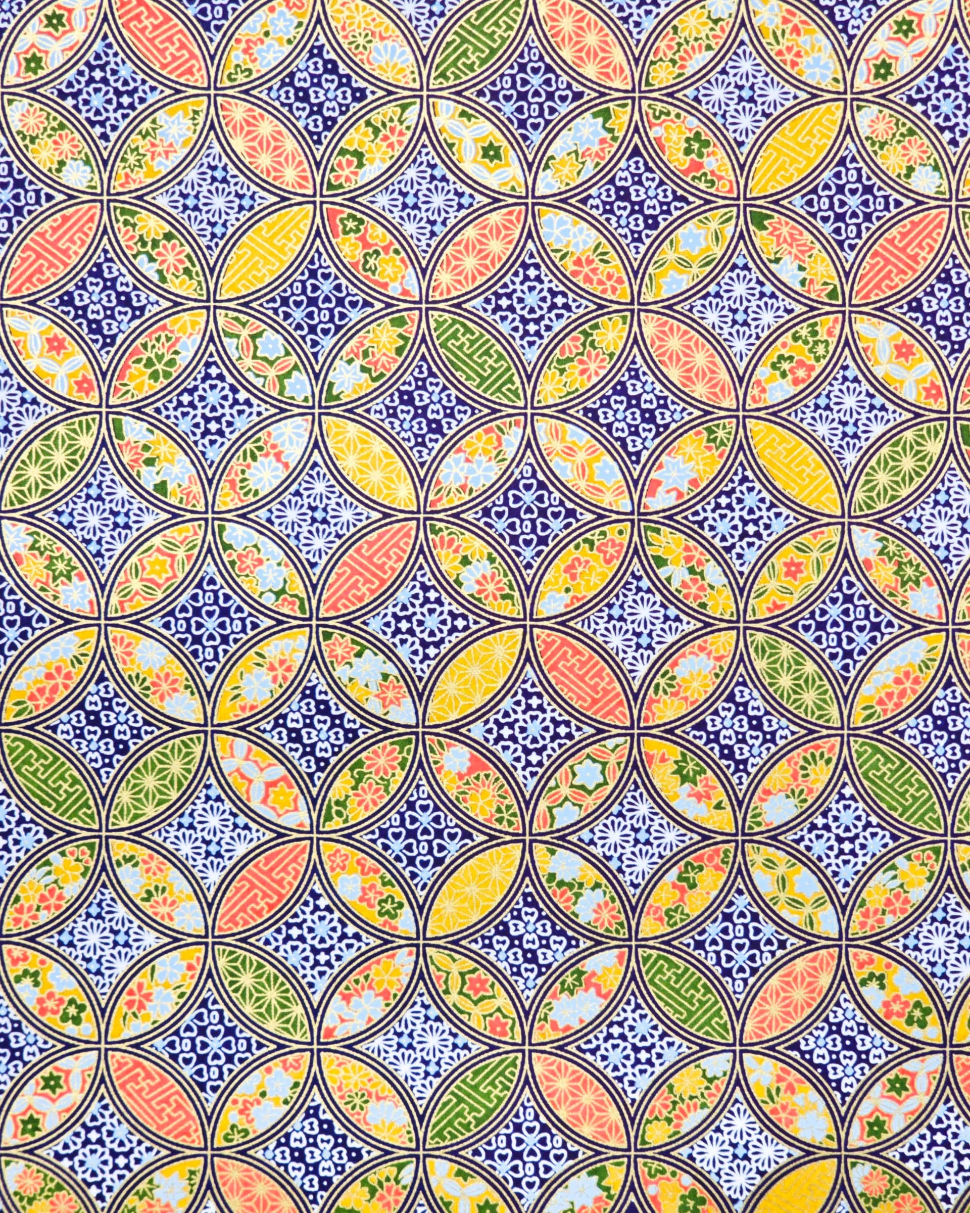 Japanese chiyogami paper featuring an orange, yellow, and green shippo design filled with assorted floral and geometric designs on a dark blue background with metallic gold details. The centers of the shippos features assorted white floral outlines.