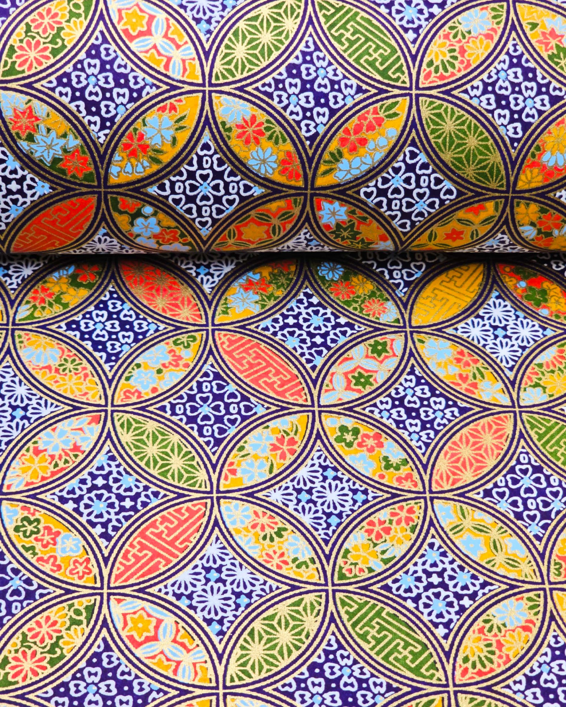 Japanese chiyogami paper featuring an orange, yellow, and green shippo design filled with assorted floral and geometric designs on a dark blue background with metallic gold details. The centers of the shippos features assorted white floral outlines.
