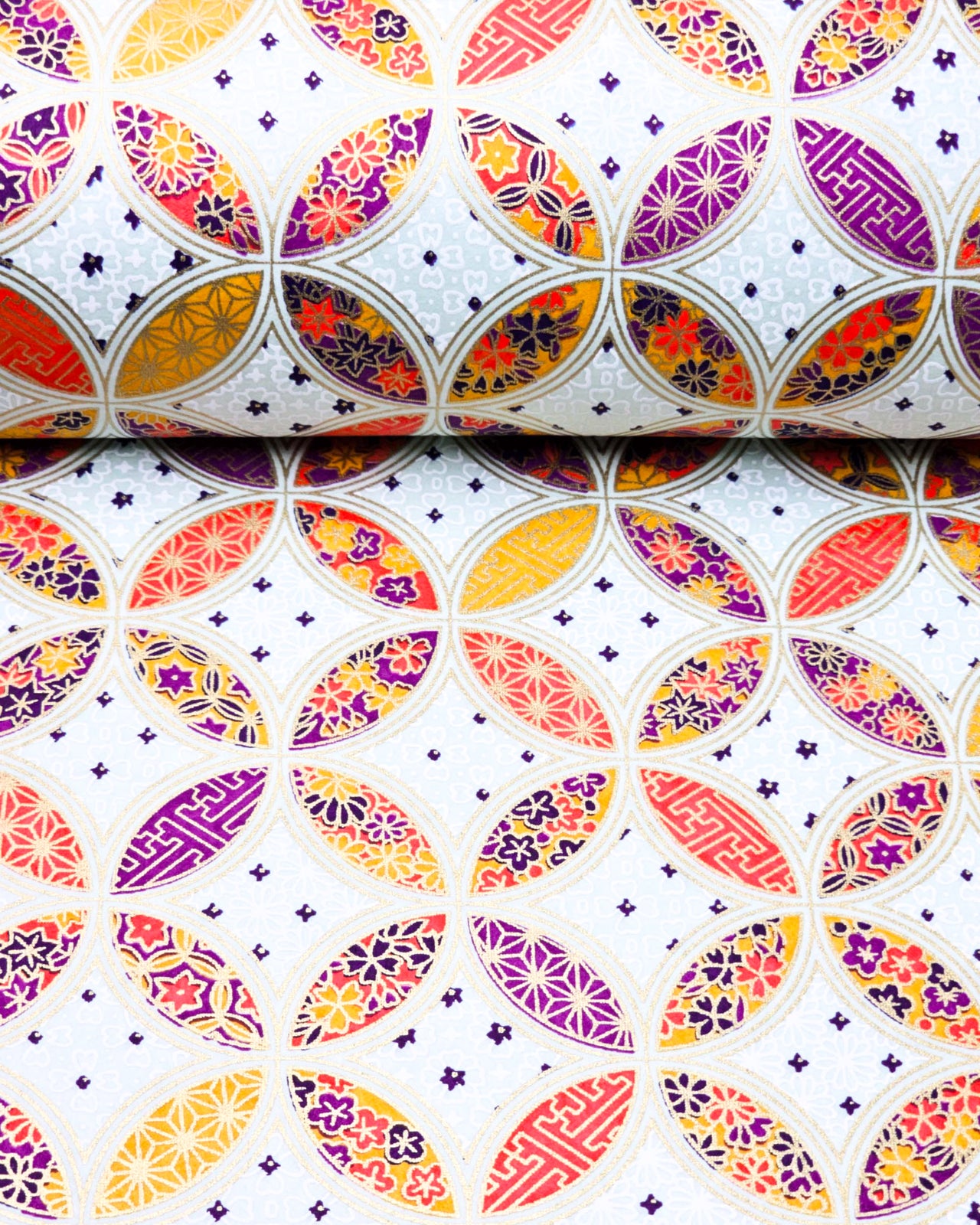 Japanese chiyogami paper featuring an orange, yellow, and purple shippo design filled with assorted floral and geometric designs on a light blue background with metallic gold details. The centers of the shippos features assorted white floral outlines.