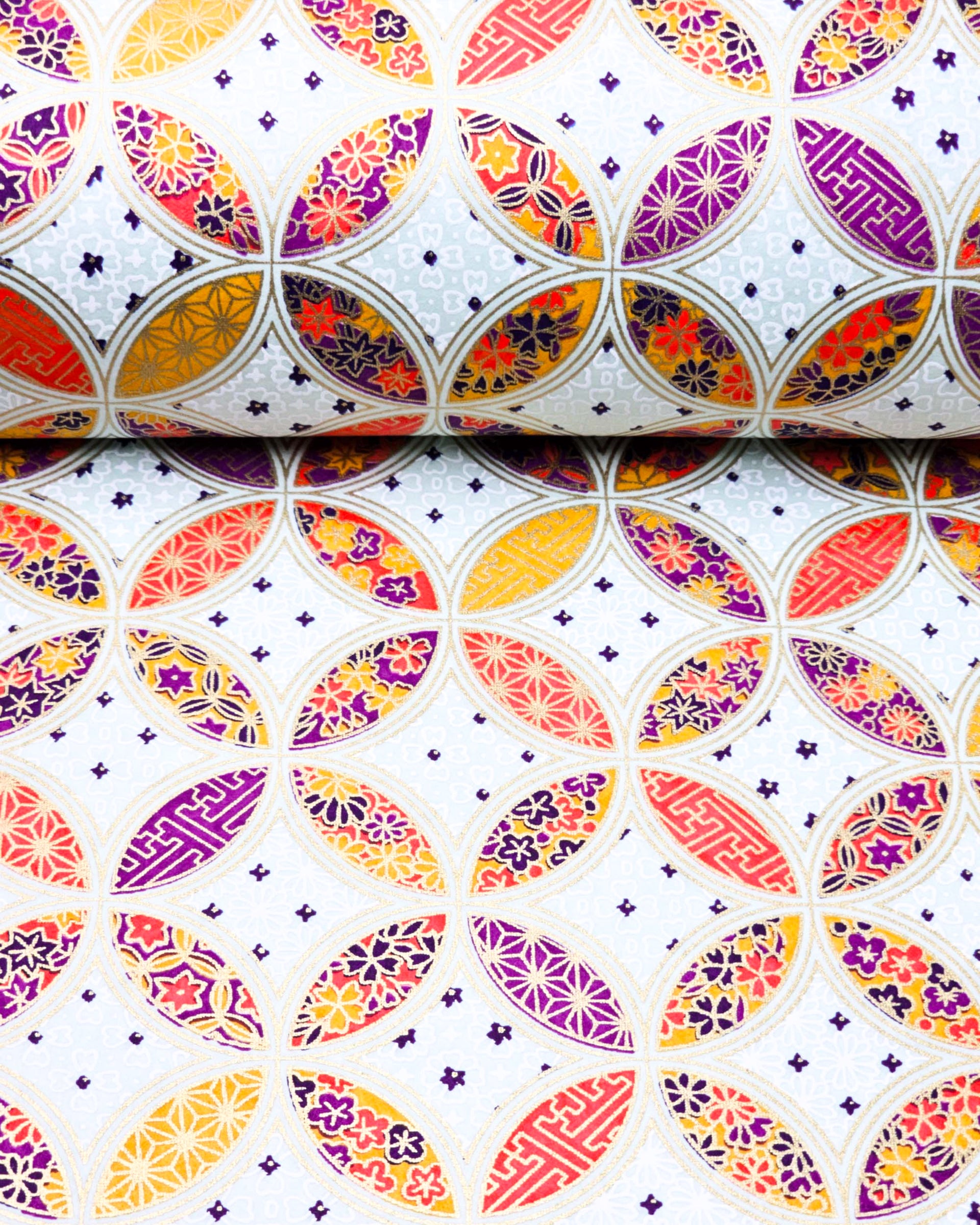 Japanese chiyogami paper featuring an orange, yellow, and purple shippo design filled with assorted floral and geometric designs on a light blue background with metallic gold details. The centers of the shippos features assorted white floral outlines.
