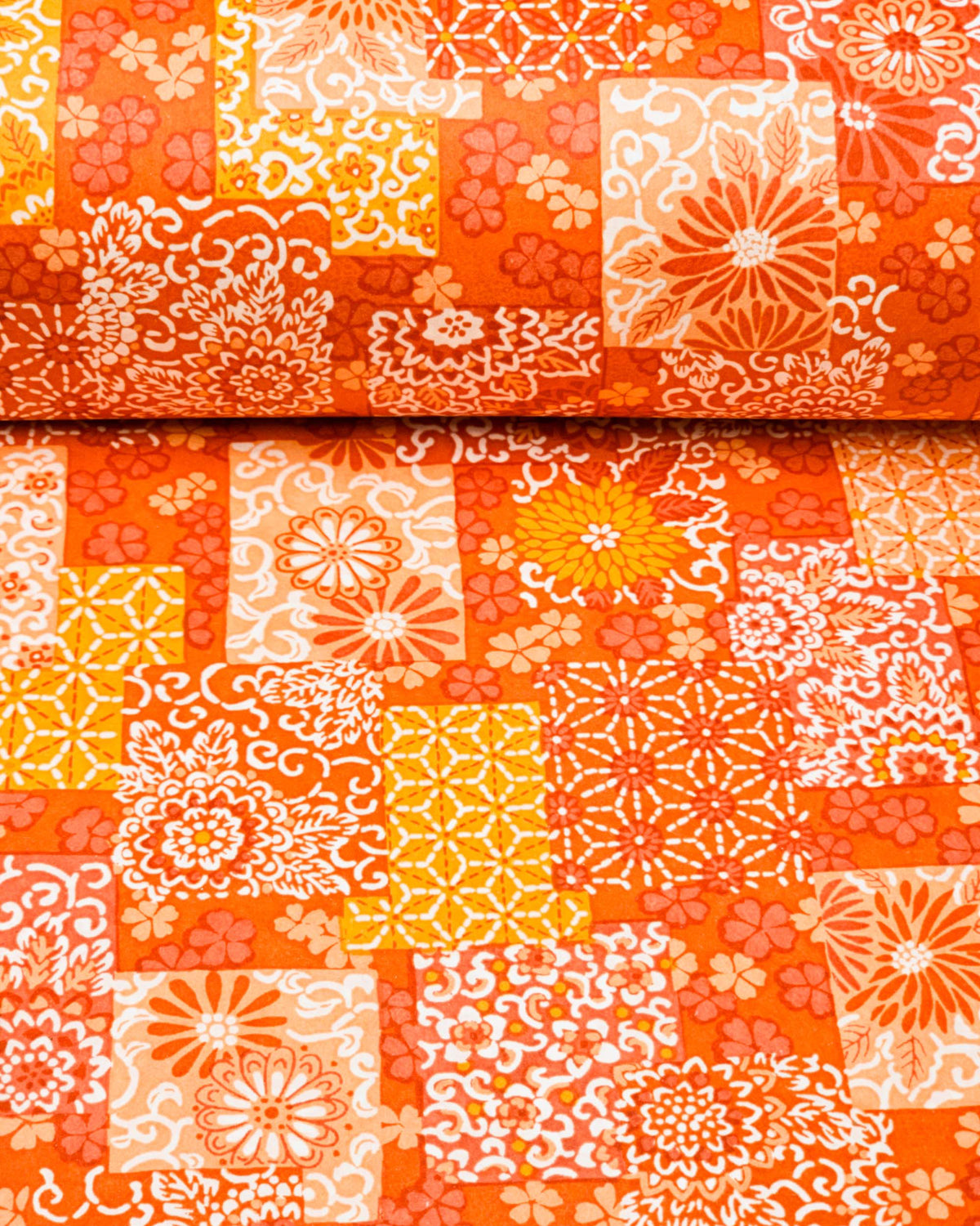 Japanese chiyogami paper featuring a patchwork-style arrangement of assorted floral and traditional Japanese patterns in various shades of orange and white, shown rolled to highlight the design.