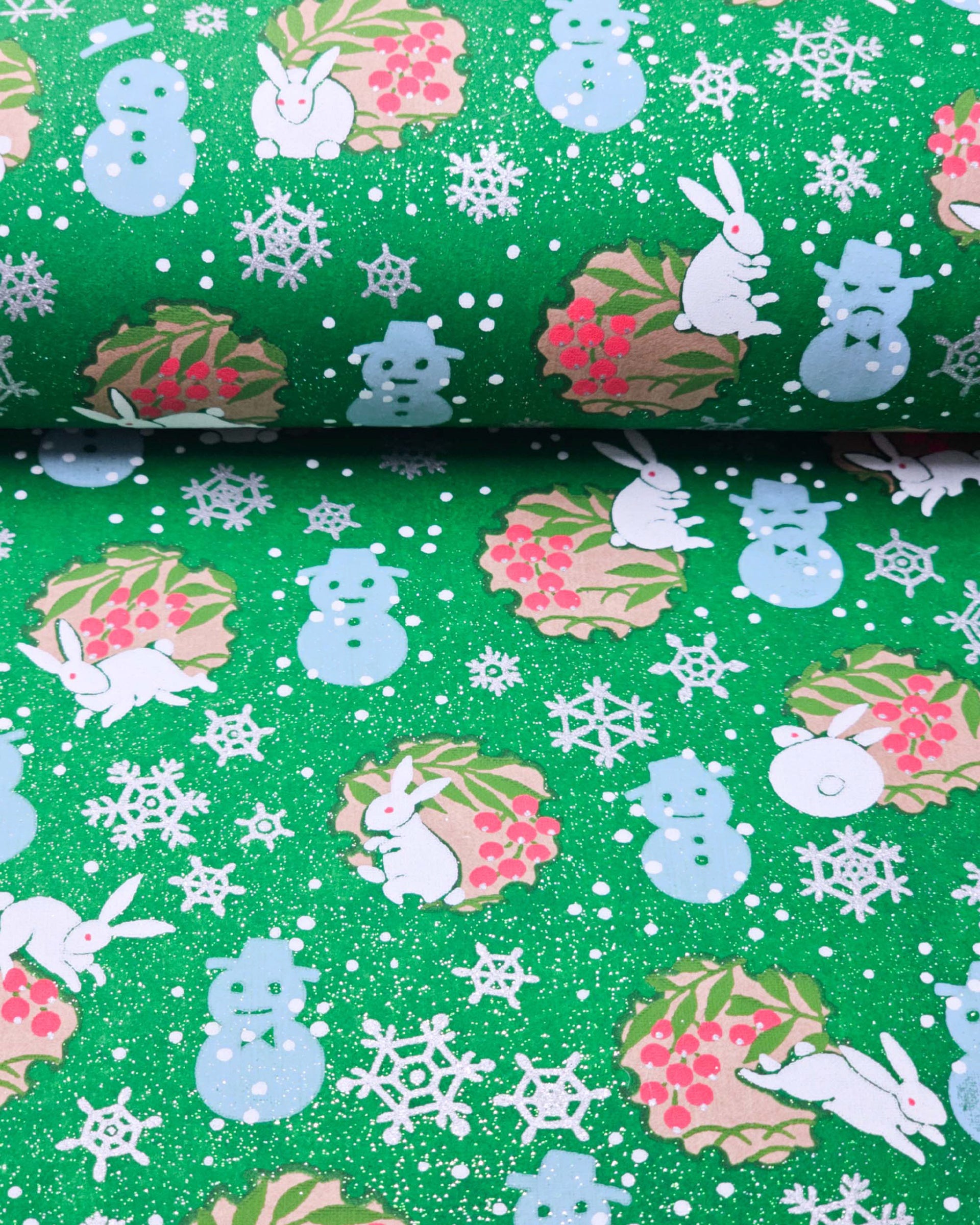 Japanese chiyogami paper featuring light blue snowmen, white rabbits, silver snowflakes, and brown yukiwas with red berries inside on a glitter green background.