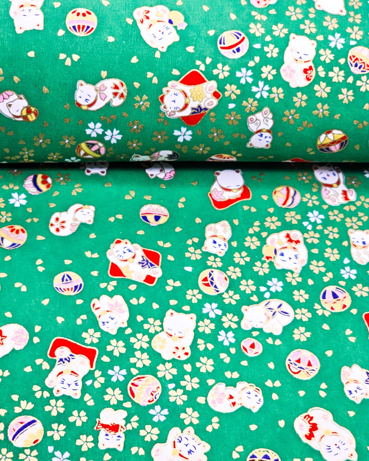 Japanese chiyogami paper featuring assorted white and red Maneki Neko (lucky cats) with small white and gold cherry blossoms and temari balls on a green background with metallic gold details.